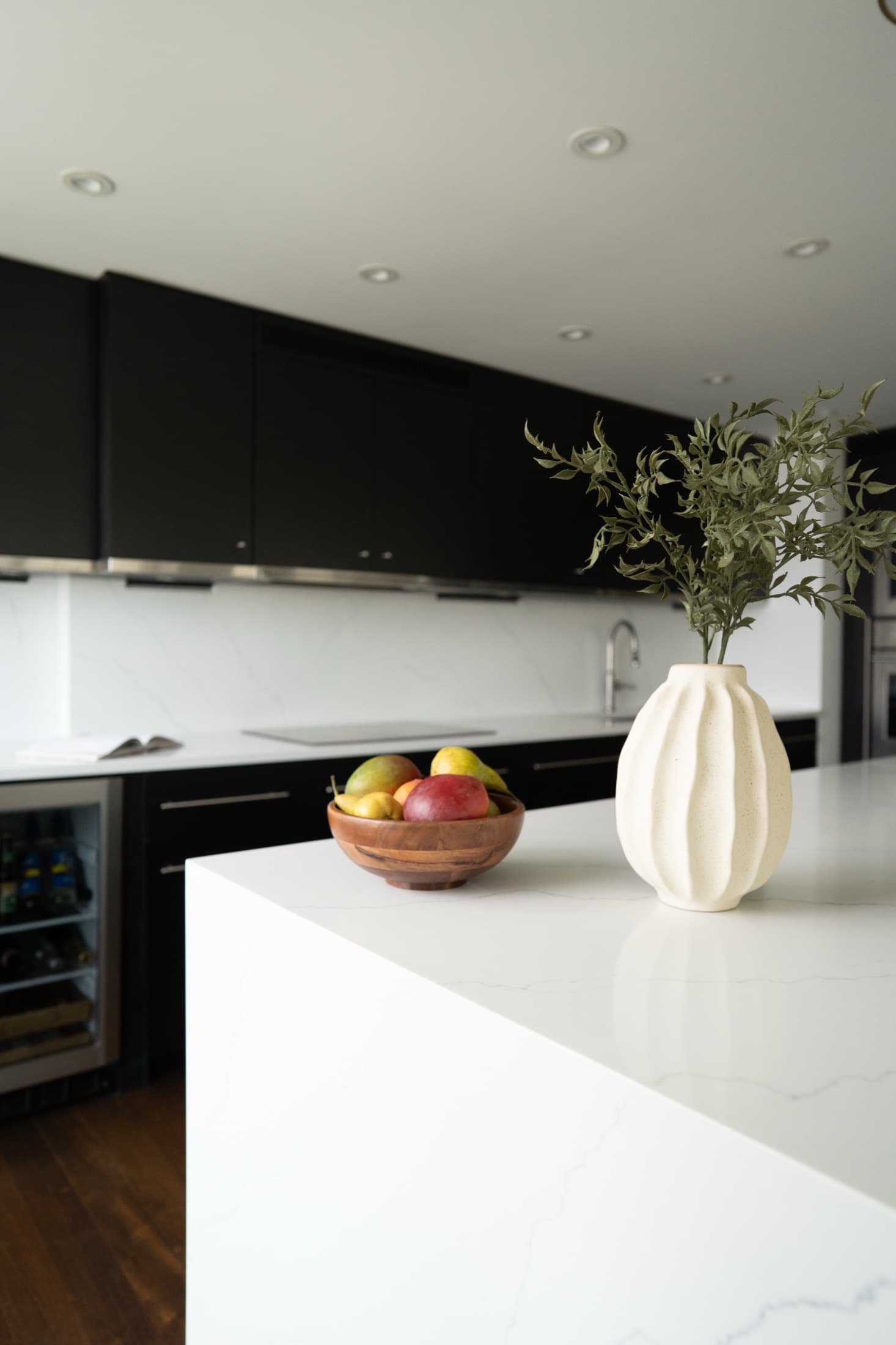 White waterfall-quartz island styled with a fluted vase of olive branches and a fruit bowl, matte-black cabinets and a wine fridge beyond.
