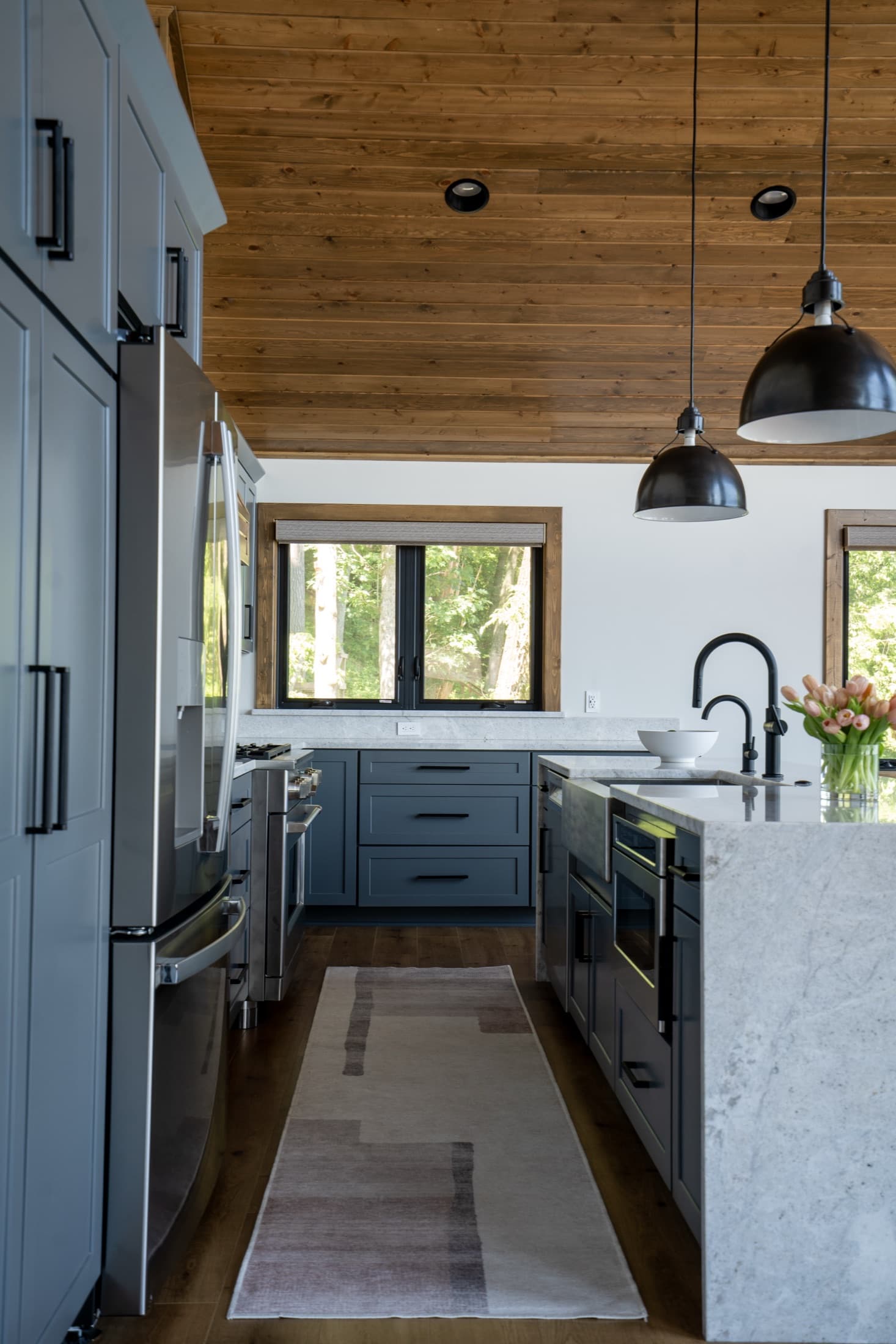 Galley view of the slate-blue lake house kitchen with a stainless refrigerator, marble island, and a runner rug on wide-plank floors.