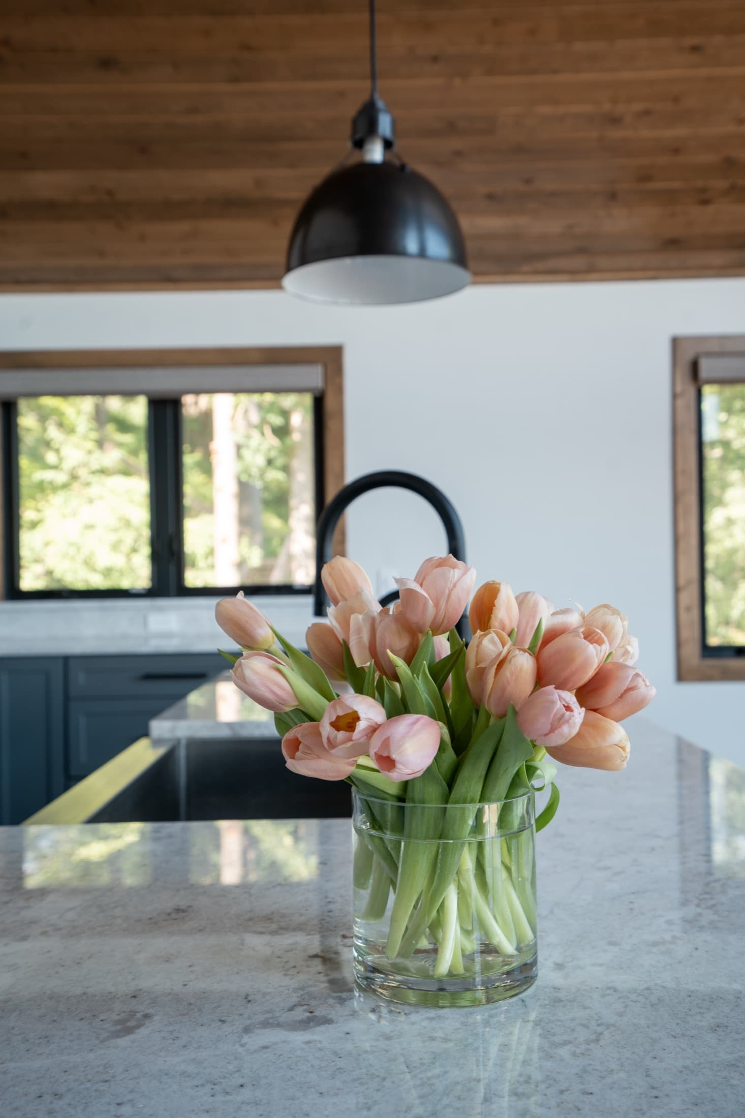Peach tulips on a marble kitchen island in a lake house with slate-blue cabinets and a warm wood ceiling.