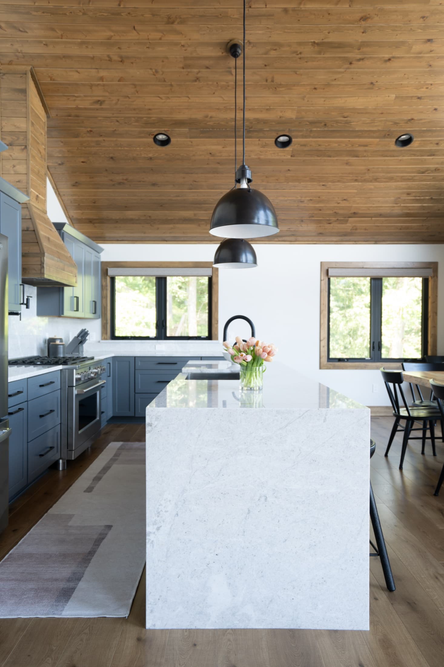 Lake house kitchen with a waterfall Carrara marble island, slate-blue cabinetry, warm wood plank ceiling, and black dome pendants.
