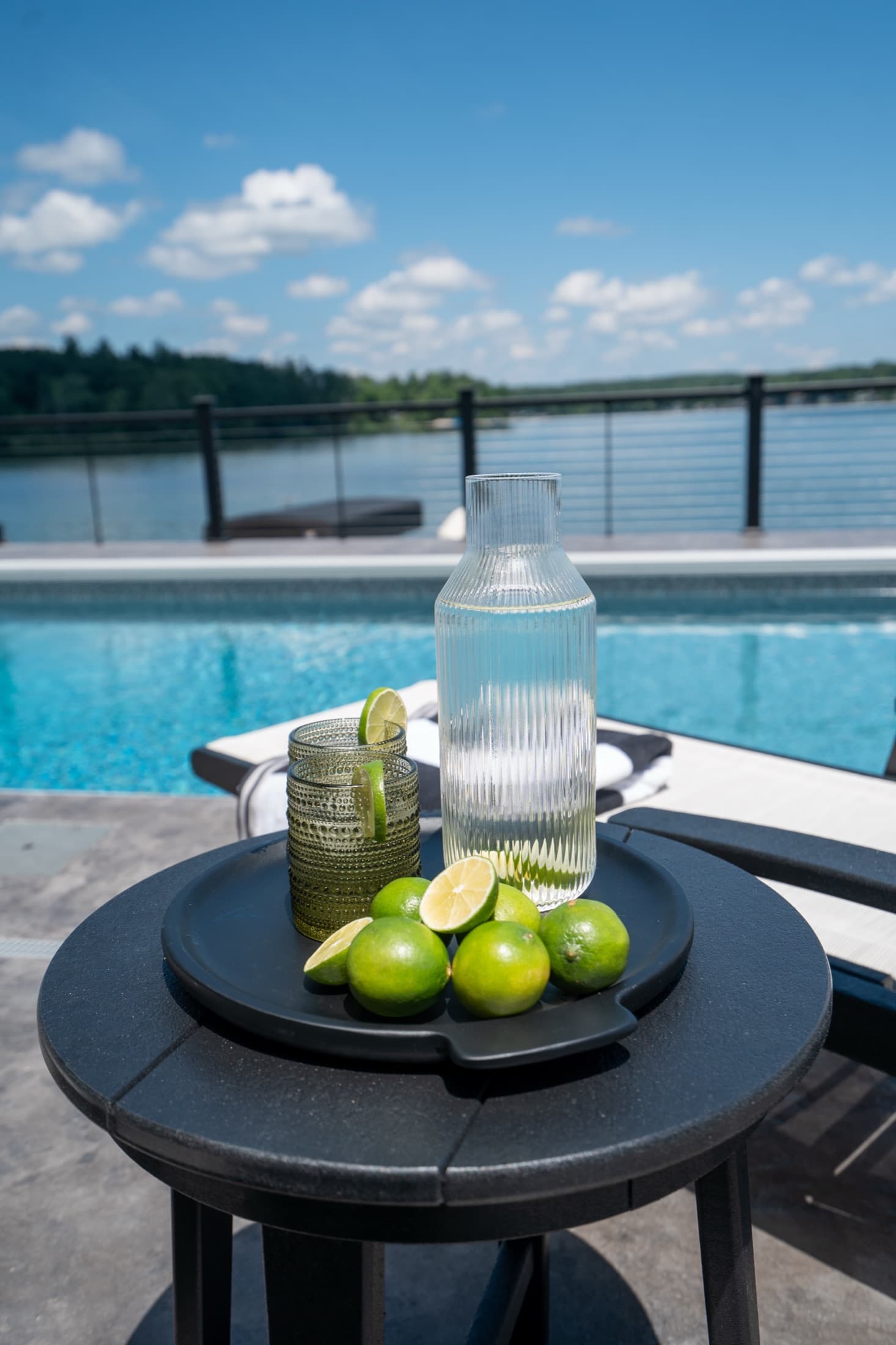 Lakeside pool deck with a black side table of drinks and limes, the blue pool and lake beyond under a summer sky.