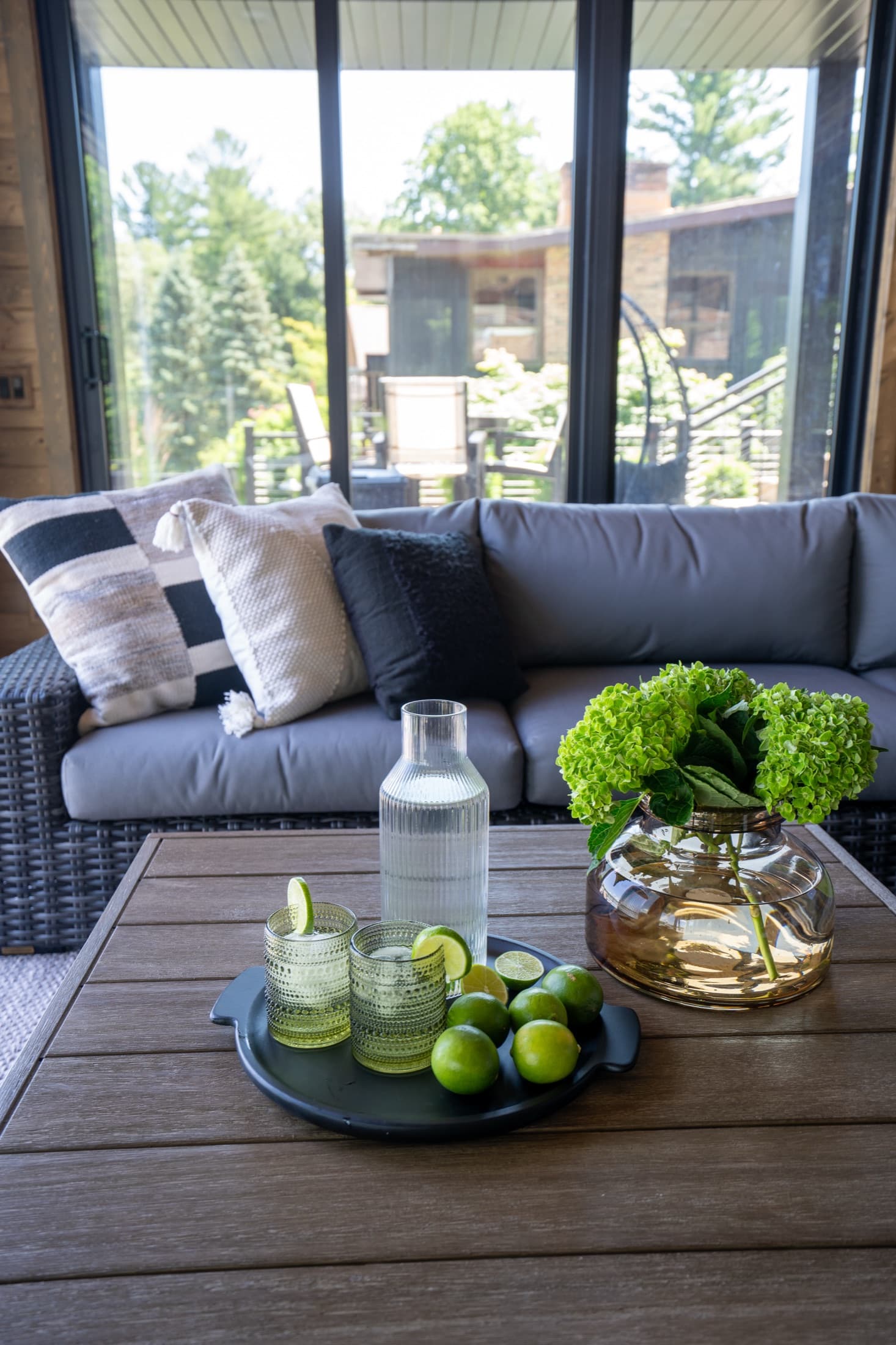 Screened porch lounge with a gray wicker sectional, striped pillows, and a wood coffee table set with drinks overlooking the wooded yard.
