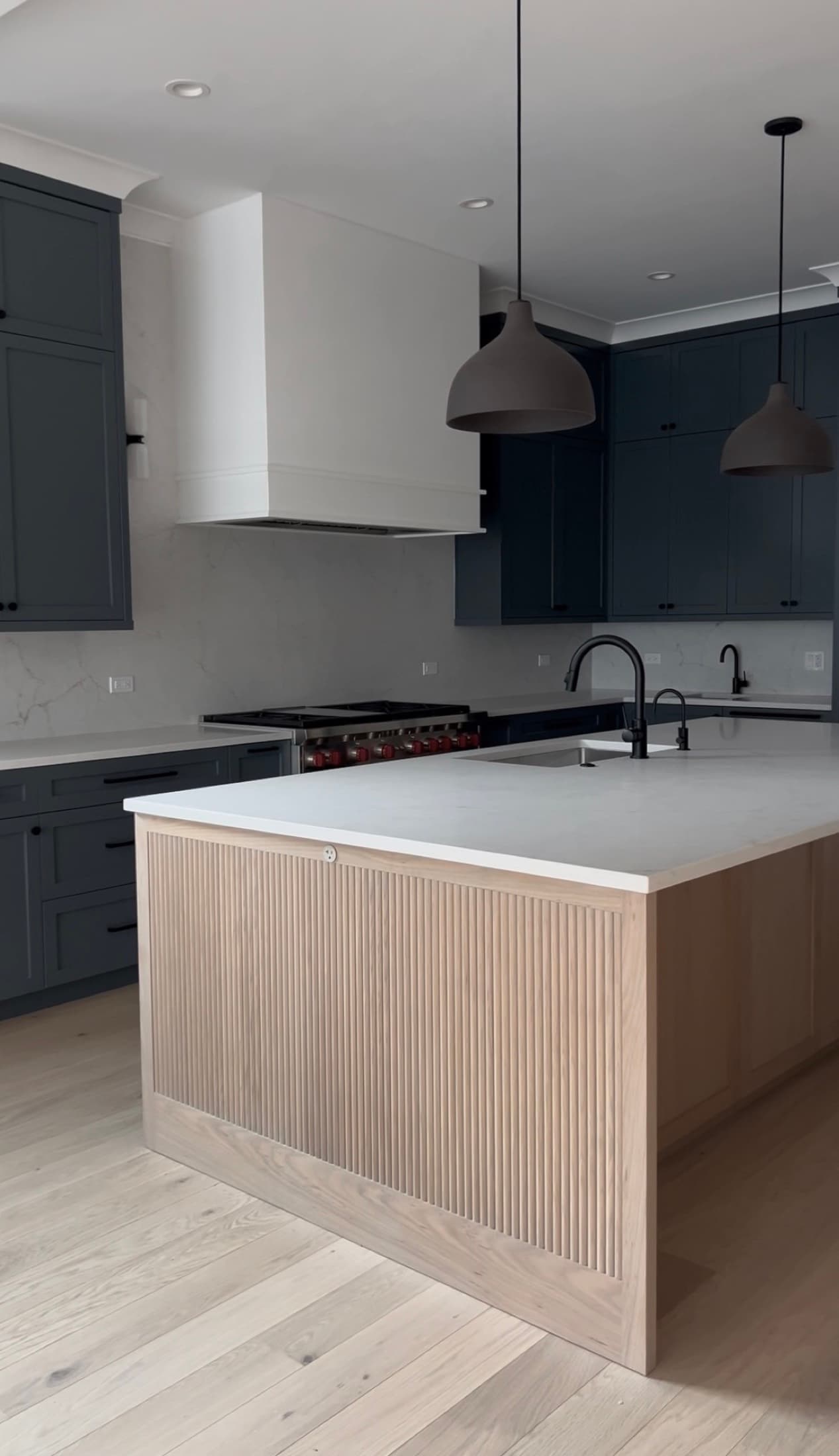 Superior Living kitchen with slate-blue cabinetry, a white-oak fluted island, marble backsplash, gas range, and black dome pendants over herringbone wood floors.
