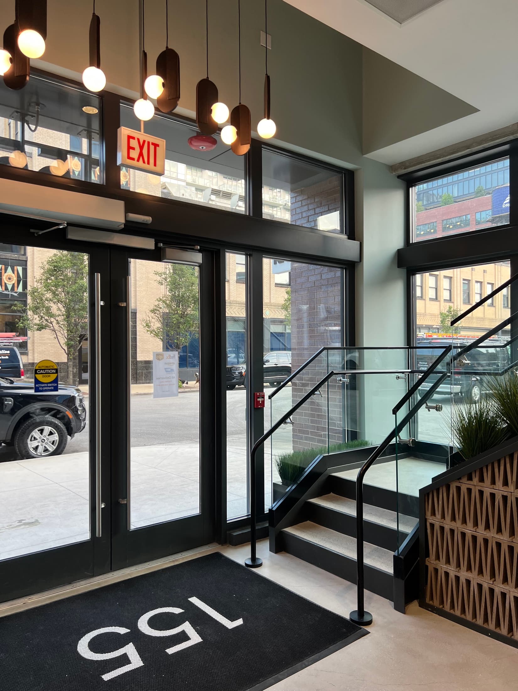 Commercial lobby entrance with black steel-framed glass doors, globe pendants, and a floating stair.